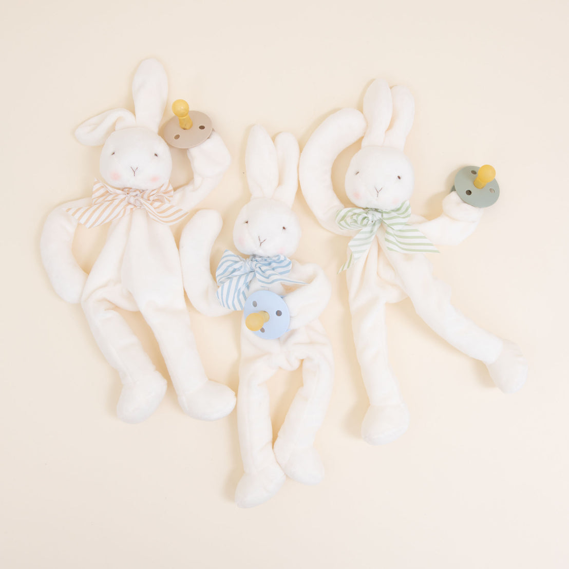 Flat lay photo of three Theodore Bunny Buddy Pacifier Holders. Stuffed animal floppy bunny made from a soft velour. Available colors include blue, green, and tan/white.