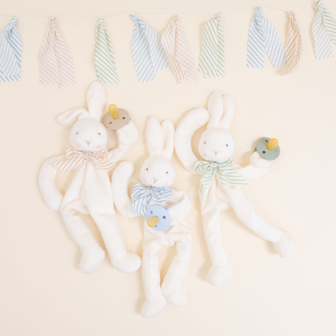 Flat lay photo of three Theodore Bunny Buddy Pacifier Holders. Stuffed animal floppy bunny made from a soft velour. Available colors include blue, green, and tan/white.