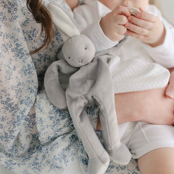 Baby boy with his mother. He is holding onto the Miles Silly Bunny Pacifier Holder, a grey bunny stuffed animal