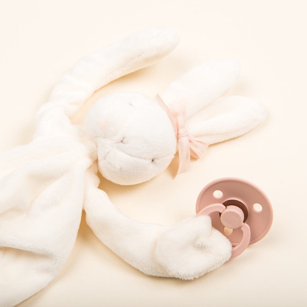 The Silly Bunny Buddy, featuring a soft and plush design with floppy ears and a pink ribbon around its neck, rests on a light background next to an accompanying pink baby pacifier, making it the perfect baby gift.