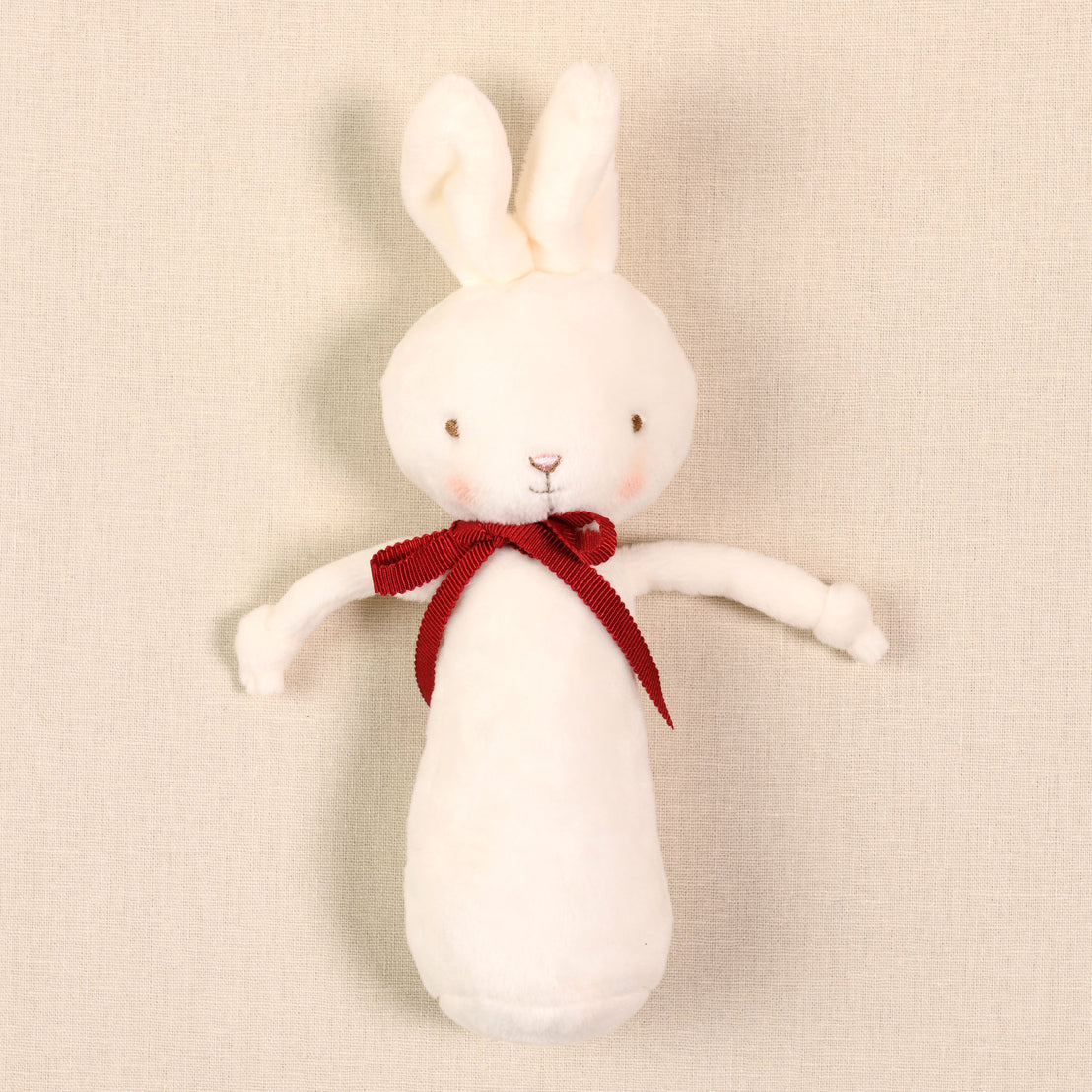 The White Bunny Chime Rattle is an adorable plush rabbit adorned with a red bow, featuring charming stitched facial details and slightly blushed cheeks against its velour cream surface. Baby safe, it adds a whimsical touch while emitting nostalgic noise with its gentle crinkling.