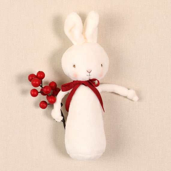 The White Bunny Chime Rattle is a charming soft velour toy embellished with a red ribbon, holding a sprig of red berries against a light beige background. It is both adorable and safe for babies to enjoy.