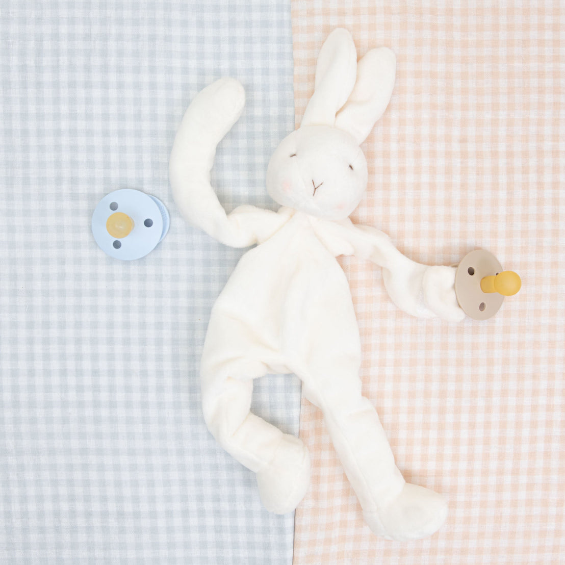 An Isla Silly Bunny Buddy on a contrasting pastel checkered background. The silly bunny is holding onto a pacifier in vanilla and another pacifier in blue is placed opposite to it. 