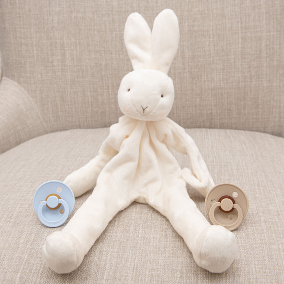 An Isla Silly Bunny Buddy sits on a beige sofa, flanked by two pacifiers, one blue and one vanilla, placed on either side of it.