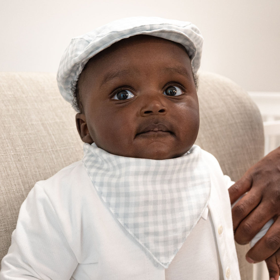 A baby dressed in a white outfit with a blue Isla Bandana Bib and matching checkered newsboy cap looks curiously at the camera.