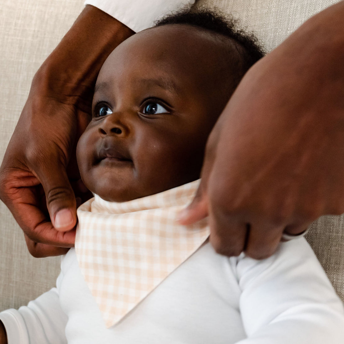 A close-up of a newborn wearing a tan Isla Bandana Bib, focusing on the baby's face looking upwards and framed by an adult's arms.