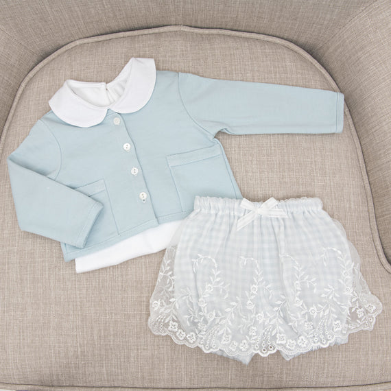 A light blue Isla French Terry Jacket with a white collared onesie paired with the matching Isla Bubble Skirt with lace embroidery placed on a textured beige chair.