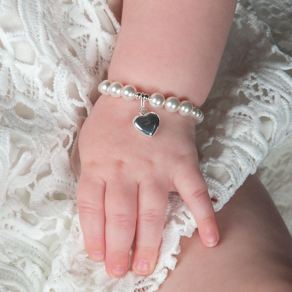 A baby's hand rests on white lace fabric, adorned with the Silver Heart Charm Bracelet & Gift Box, featuring a dazzling pearl and a sterling silver heart charm—an ideal baby shower gift.