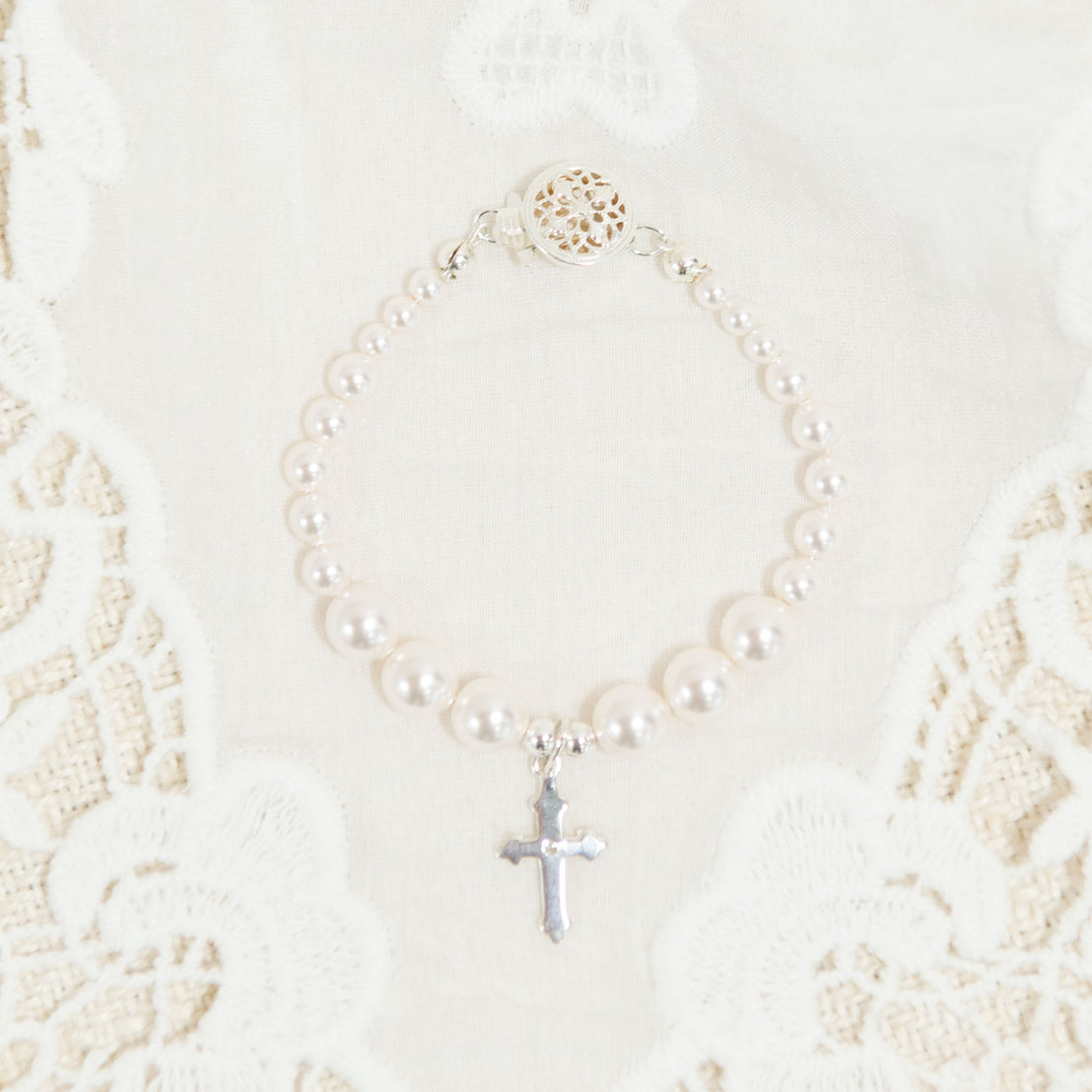 A flat lay photo baby jewelry: the white luster pearl bracelet with a silver cross in the middle. Photographed on top of the Adeline Christening Gown.