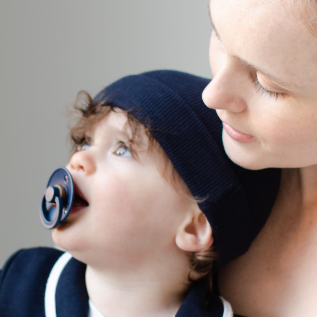 A mother gently holds her baby, who is looking upwards with a Bibs Pacifier in Deep Space in mouth. The baby wears a matching navy blue hats, against a soft gray background.