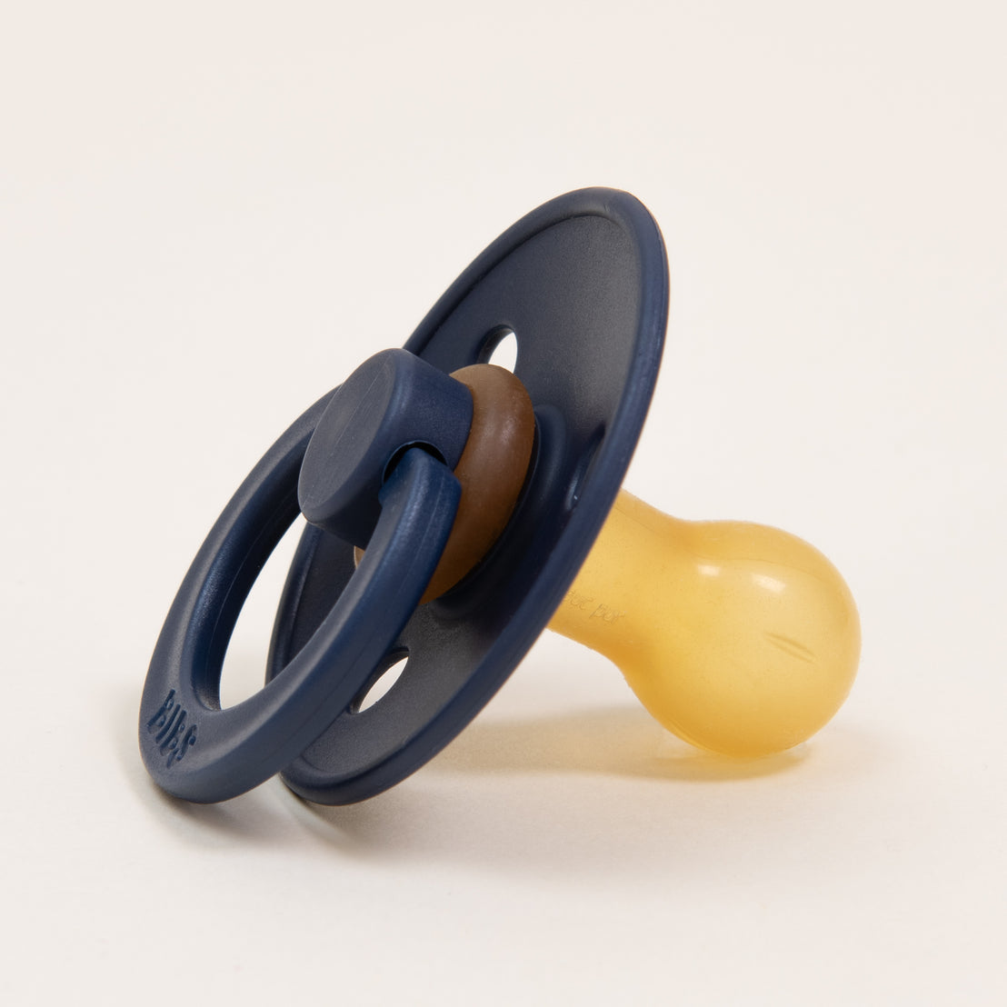 A close-up image of a blue and brown Bibs Pacifier in Deep Space with a natural rubber nipple, resting on a light beige background.