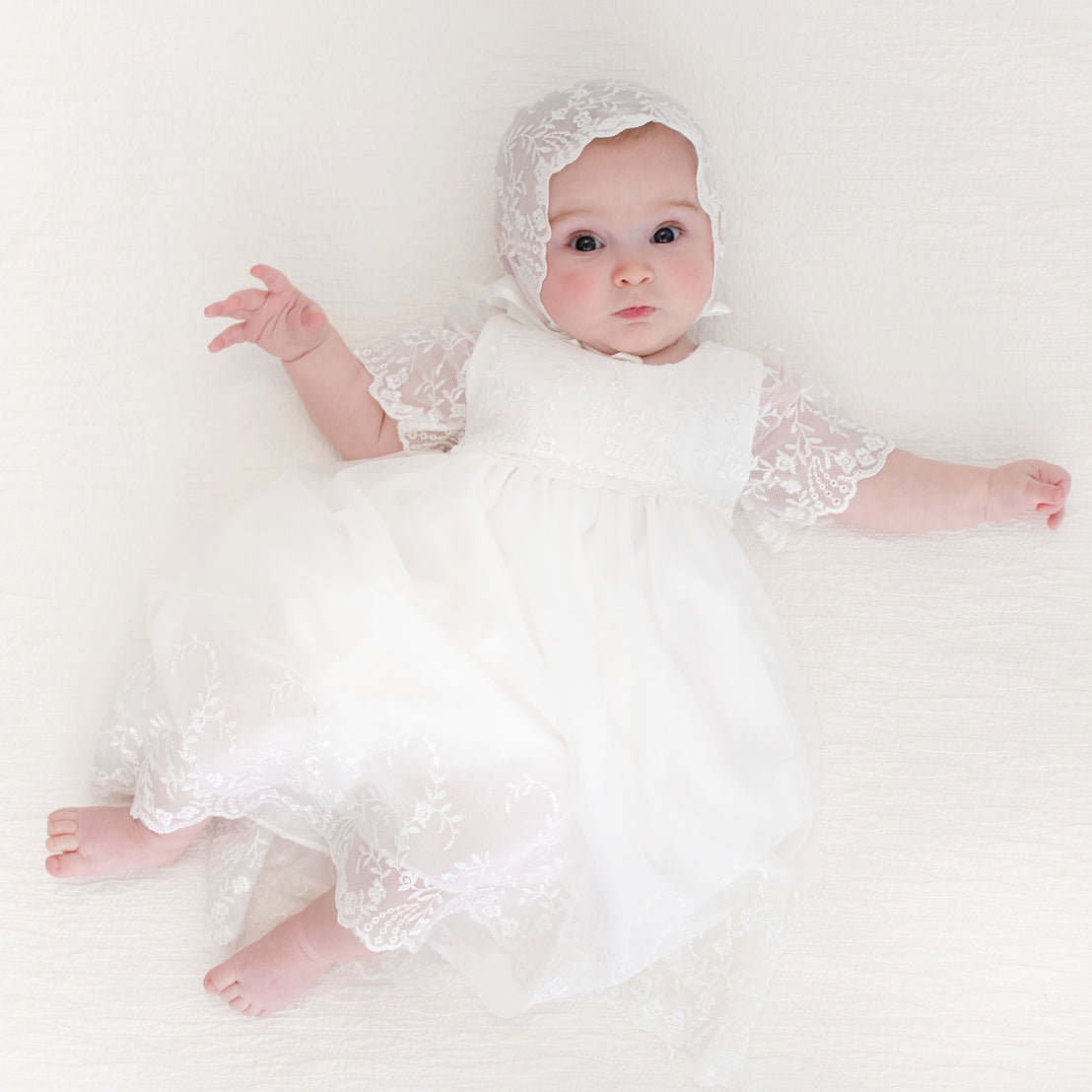 Baby girl dressed in the Ella Romper Dress and Ella Lace Bonnet, gazing curiously at the camera with open arms.