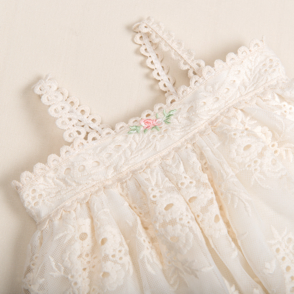 Close-up of a delicate white lace Clementine Dress & Bloomers with a focus on the detailed stitching, tiny pink and green floral embroidery, and scalloped edges.