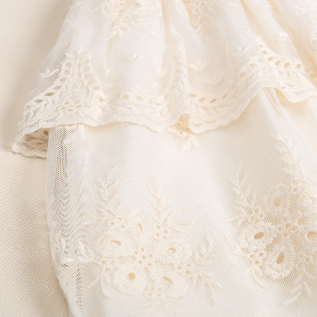 Close-up of Clementine Dress & Bloomers, an upscale, vintage ivory lace fabric with floral embroidery and scalloped edges, displaying intricate designs and delicate needlework, perfect for baptism attire.