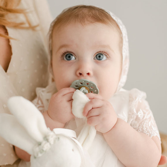 A newborn baby girl with a sage pacifier in her mouth, part of the Ella Pacifier Set that includes Sage and Ivory colors.