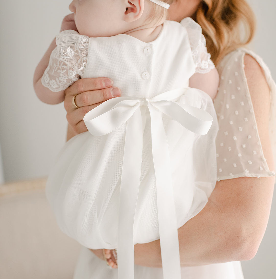 A photo showcasing the back of the Ella Christening Gown, worn by a baby girl who is held by her mother.