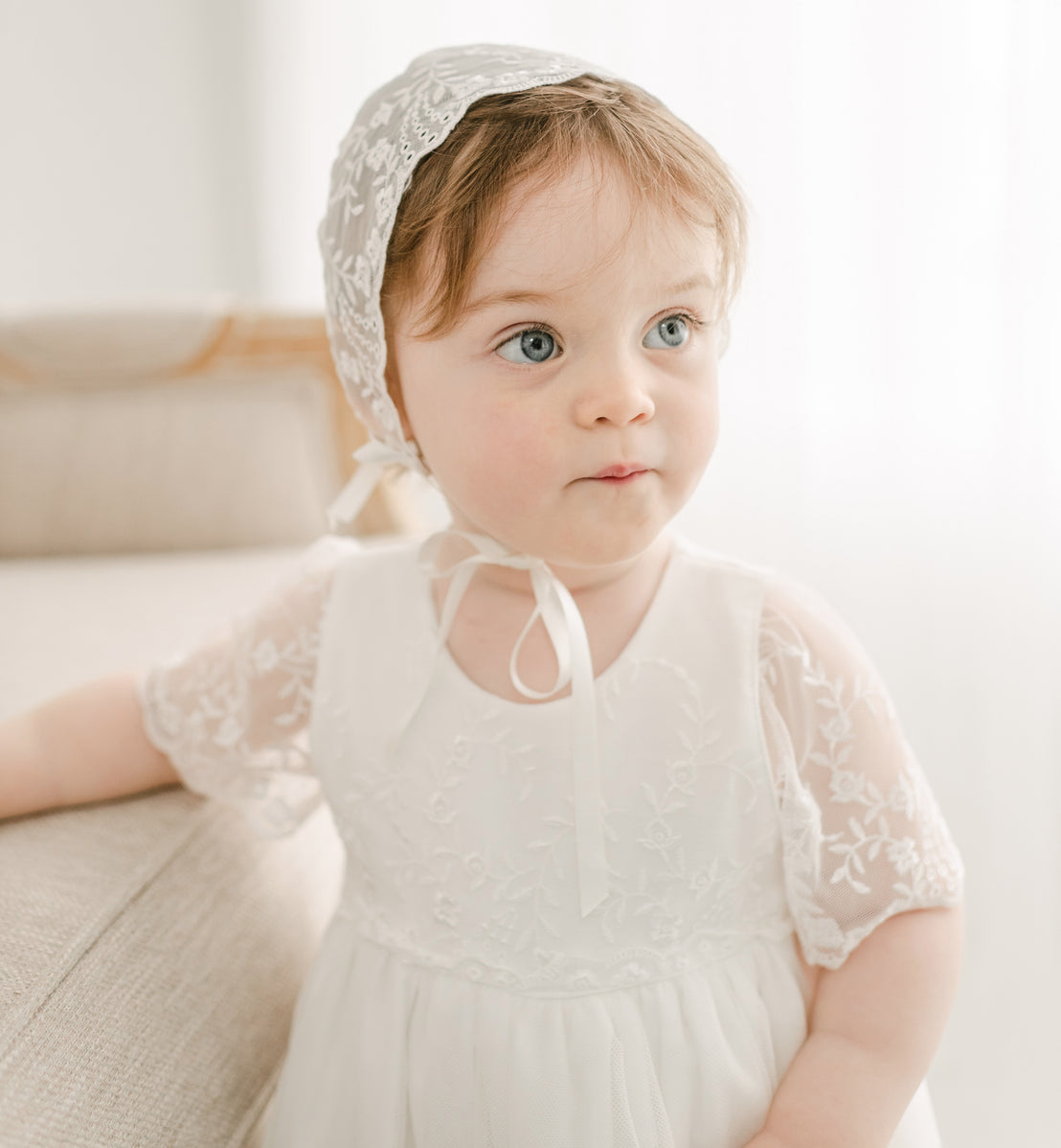 A baby girl with red hair wearing the Ella lace bonnet and full-length christening gown.