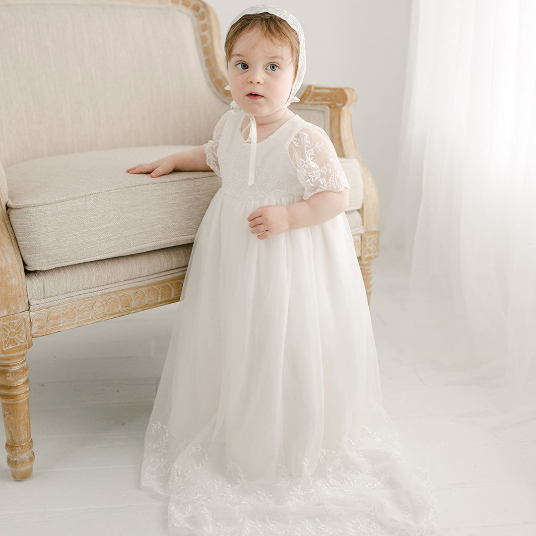 Baby girl wearing the Ella Christening Gown and standing next to a upholstered chair.