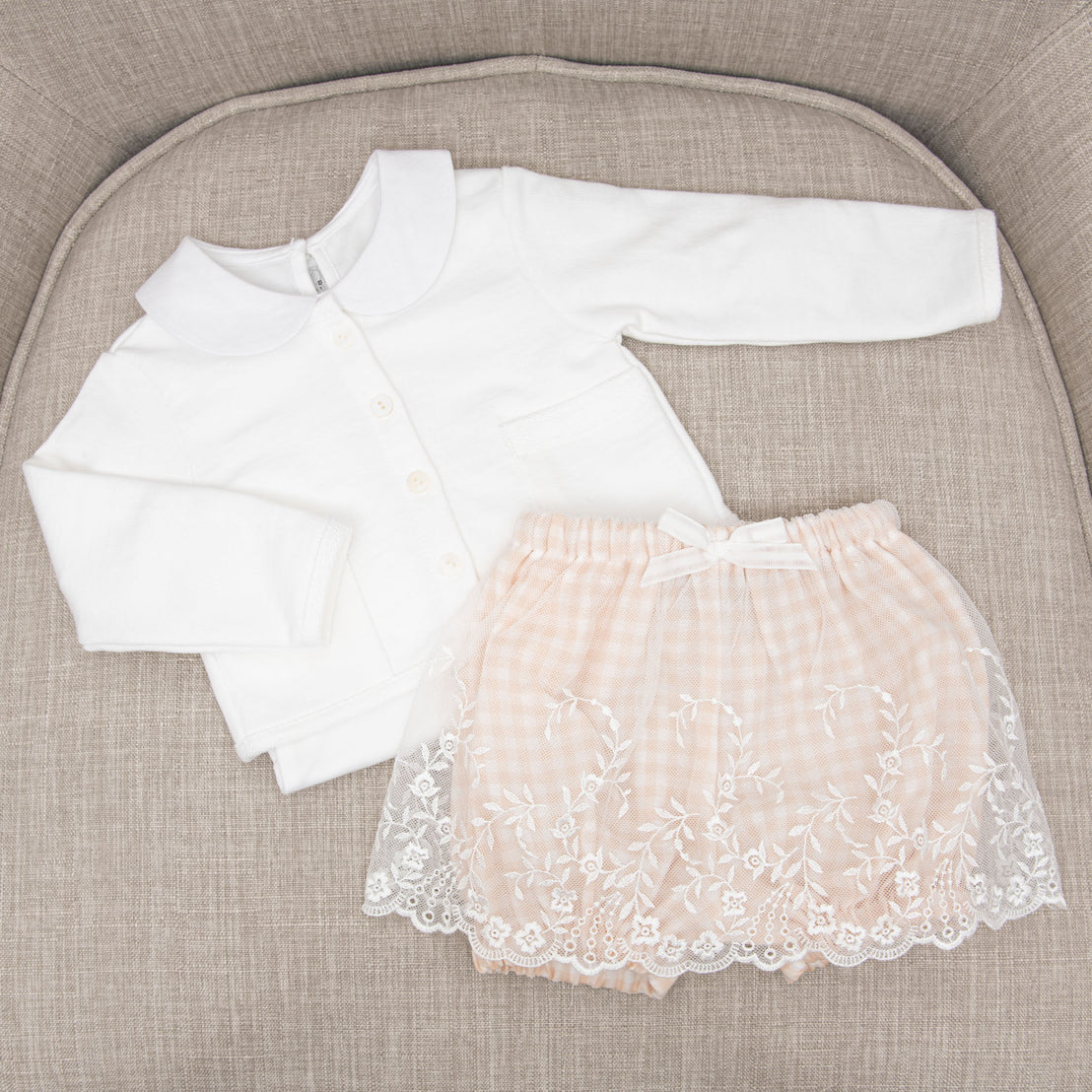 A white Isla French Terry Jacket with a white collared onesie paired with the matching peach Isla Bubble Skirt with lace embroidery placed on a textured beige chair.