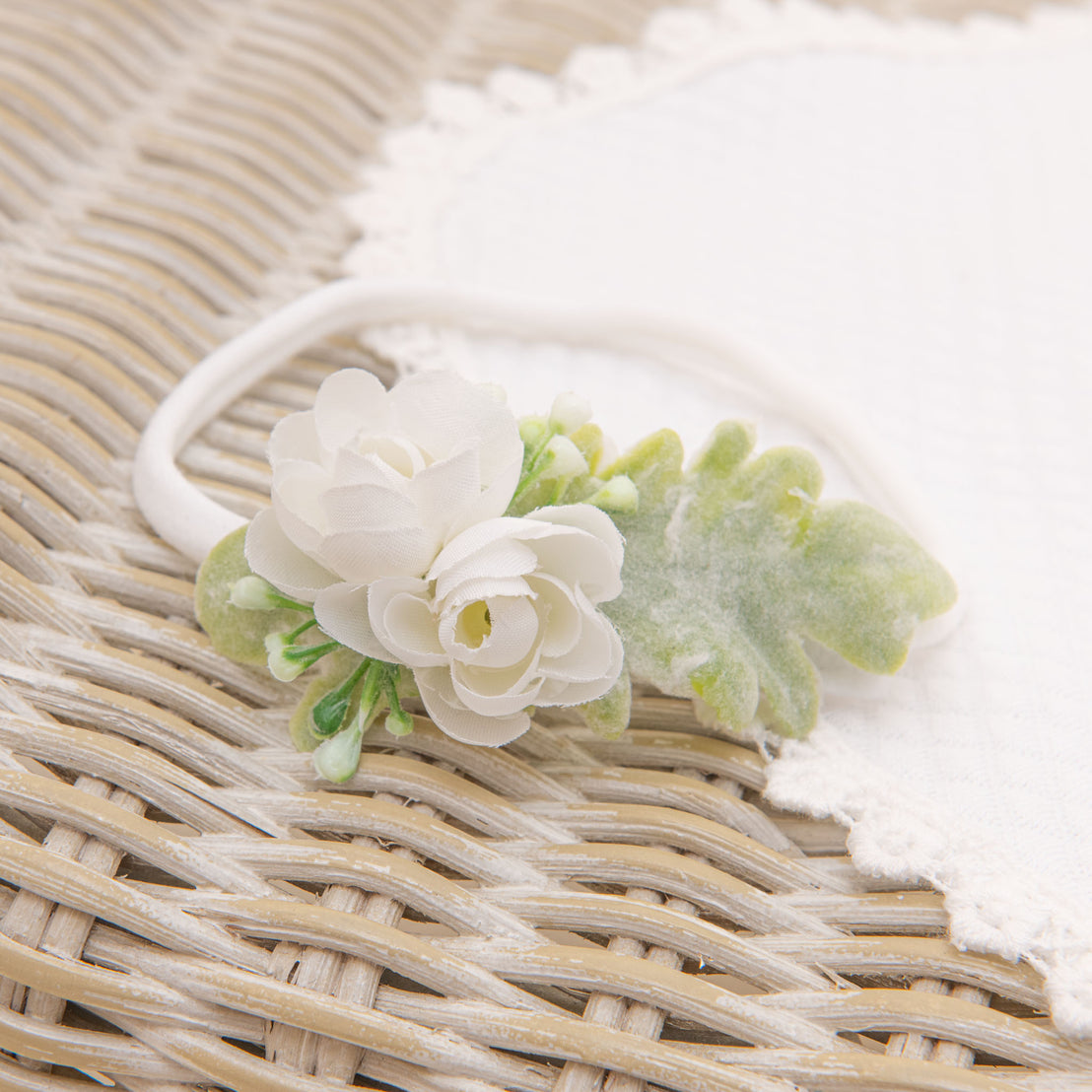 A delicate white Eliza headband with green leaves, perfect for a baptism, placed on a woven texture background.