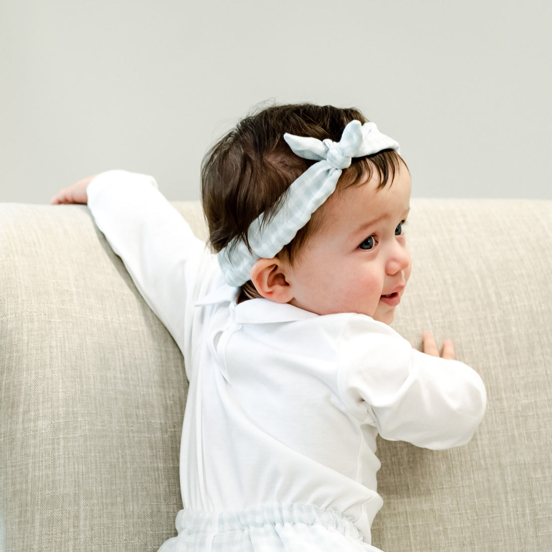 A baby girl with dark hair wearing the Isla Tie Headband and white outfit looks over her shoulder, smiling, while lying on a beige sofa.