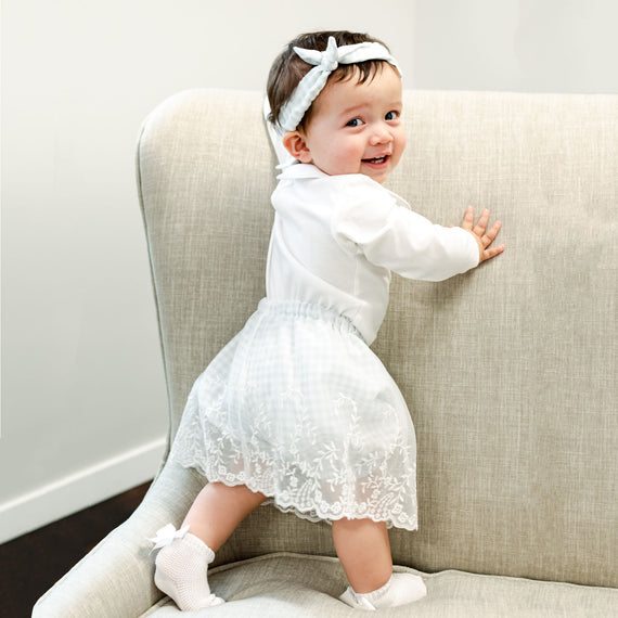 A smiling baby girl stands by holding onto a gray couch, dressed in the Isla Bubble Skirt Set and matching Tie Headband. 