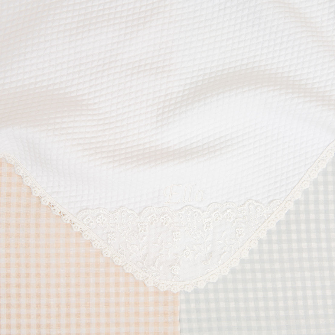 Close-up of the Isla Personalized Blanket against a backdrop of checkered blue and peach fabric. The blanket features floral embroidered lace on the corner with the name "Ella" in ivory thread.