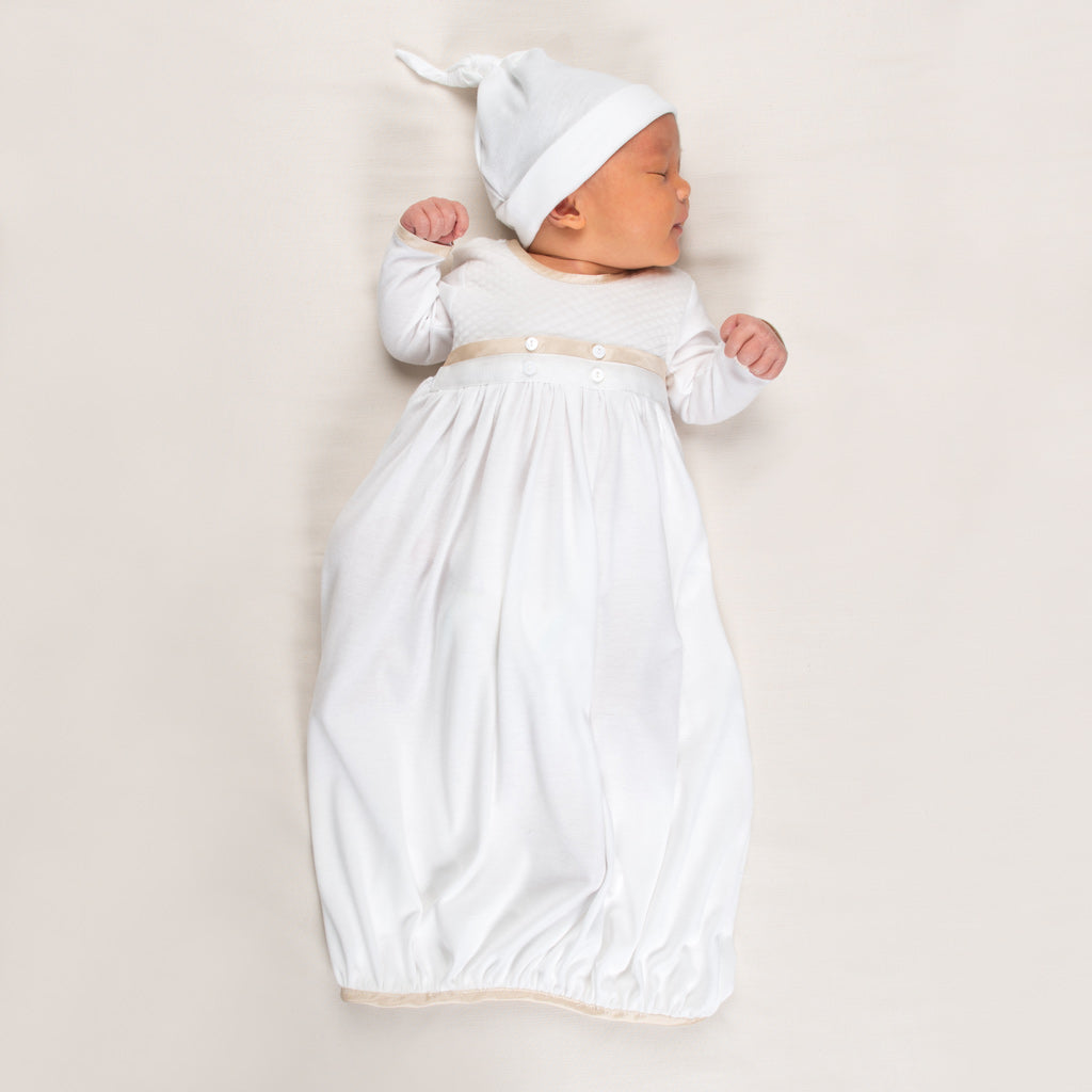 A sleeping baby dressed in a long, white pima cotton gown with buttons and the Liam Cap knotted hat lies on a soft, light-colored surface. The newborn ensemble perfectly complements the peaceful scene as the baby rests serenely on their side.