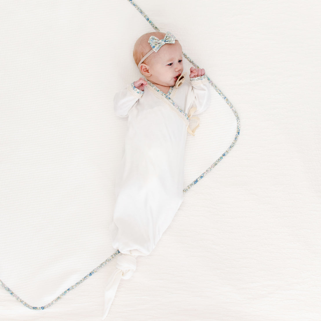 A baby dressed in a Petite Fleur Knot Gown and a bow headband lies on a soft white blanket, with a lightly colored rope loosely shaped in a heart surrounding her, creating a vintage-inspired scene.