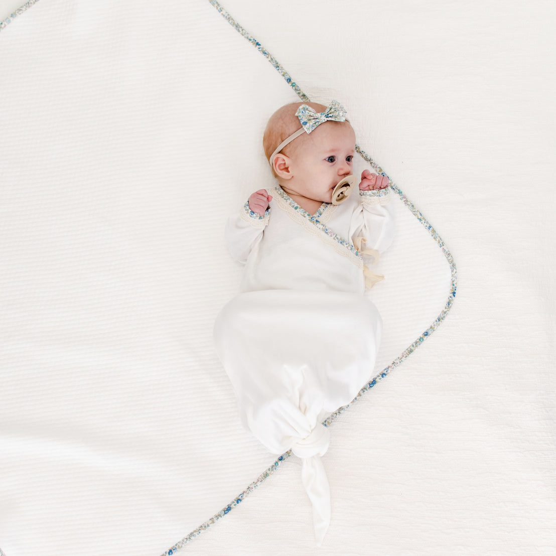 A baby in a Petite Fleur Knot Gown lies on a soft, white blanket, holding a golden pacifier and wearing a light bow headband, encircled by a delicate bluish-silver string of beads.