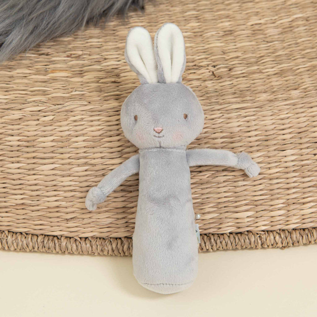 A soft, gray bunny plush toy with long ears and a minimalistic facial expression lies on a woven, circular rug. Perfect as part of an Elijah Newborn Gift Set - Save 10% or a unique baby shower gift, the bunny has a cylindrical body and short limbs.