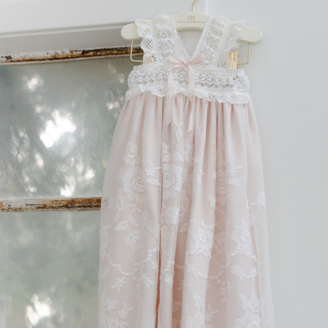 A delicate pink and white Charlotte Christening Gown  hangs on a wooden hanger against a white wall, reflecting soft light. The dress features intricate lace patterns and a flowing skirt.