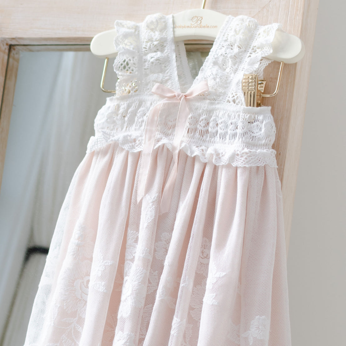 An elegant pink Charlotte Christening Gown for a little girl, featuring delicate cotton lace detailing and a satin ribbon at the chest, hanging on a wooden hanger against a soft background.