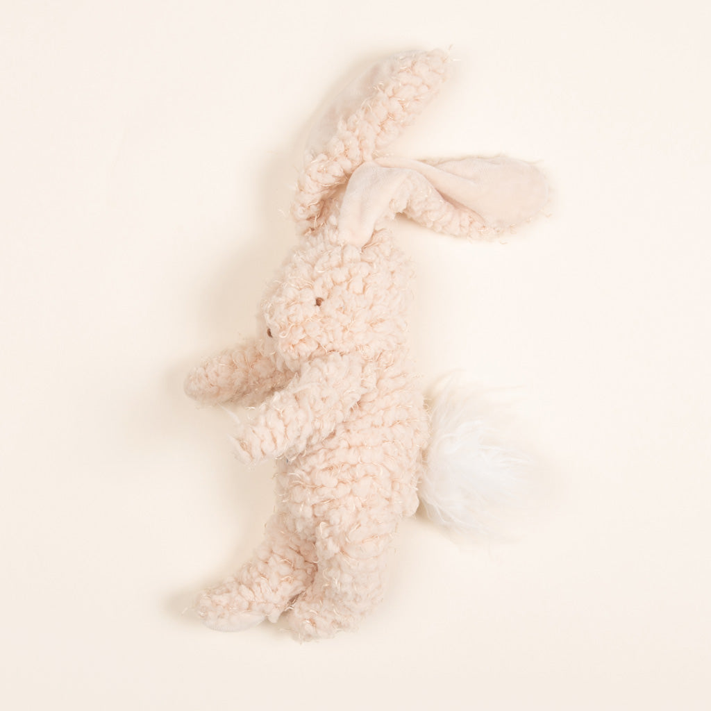 The Harey Bunny, a plush toy with long ears and a fluffy tail made from soft cream-colored fabric, is lying on its side against a plain off-white background. This adorable bunny friend makes the perfect baby gift with its irresistibly soft fur.