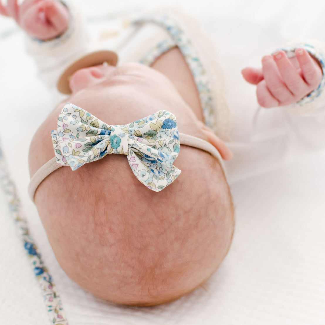 Close-up of a newborn baby's head wearing a Petite Fleur Headband, with parts of a blanket visible in a soft-focus background.