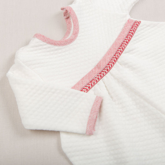 The Sweetheart Newborn Romper showcases a close-up view of its white textured design, highlighted by red trim. Made from 100% quilted cotton, this baby's Christmas ensemble includes long sleeves adorned with decorative red stitching at the neckline and an angled seam across the front. The cozy and soft fabric makes it an ideal keepsake baby Christmas outfit.