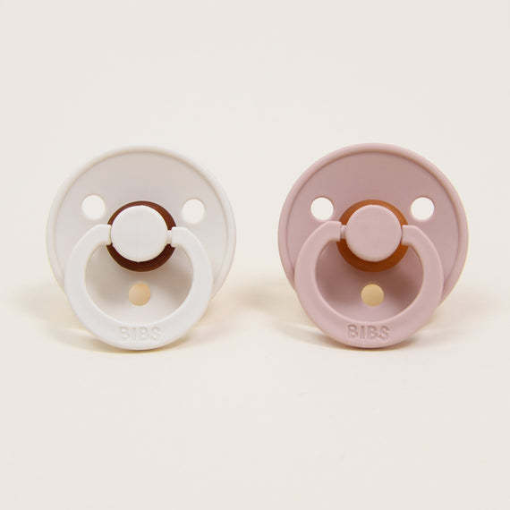 The Tessa Pacifier Set, featuring two baby pacifiers crafted with natural rubber, is displayed side by side on a neutral background. The left one is in ivory and the right in blush, each showcasing a circular base and a round handle embossed with "BIBS," a trusted Danish brand known for helping babies self-soothe.