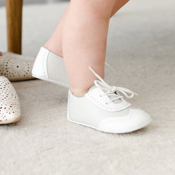 A baby wearing white Baby Beau & Belle clothing and handmade baby shoes stands next to an adult seated on a chair, who is wearing Ivory Two Tone Wingtip Shoes. The scene is set on a light-colored carpet.