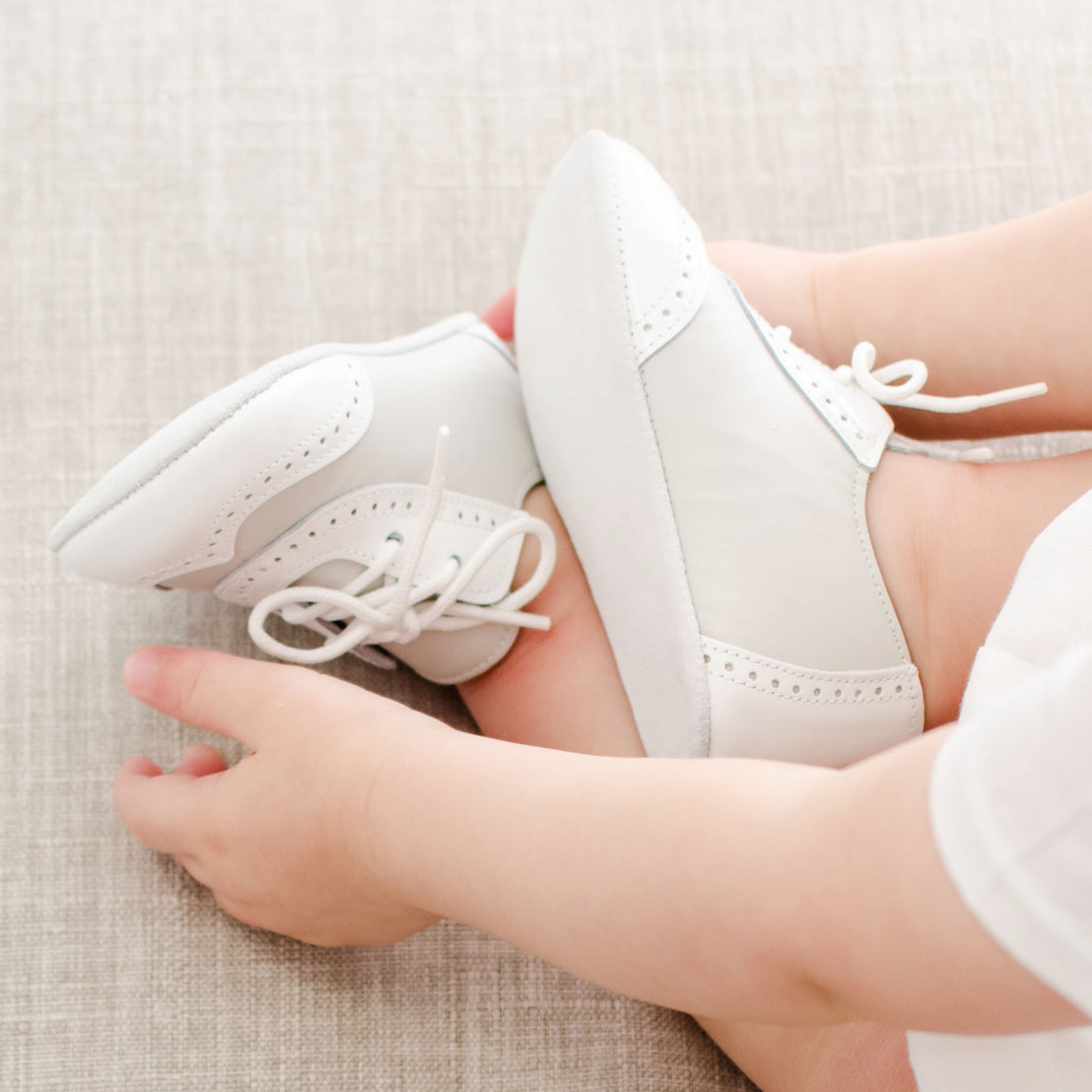 A close-up image of a baby wearing handmade leather baby shoes while sitting on a light-colored fabric surface. The baby is holding one of their feet, showcasing the tiny details of the Ivory Two Tone Wingtip Shoes with laces and decorative perforations.