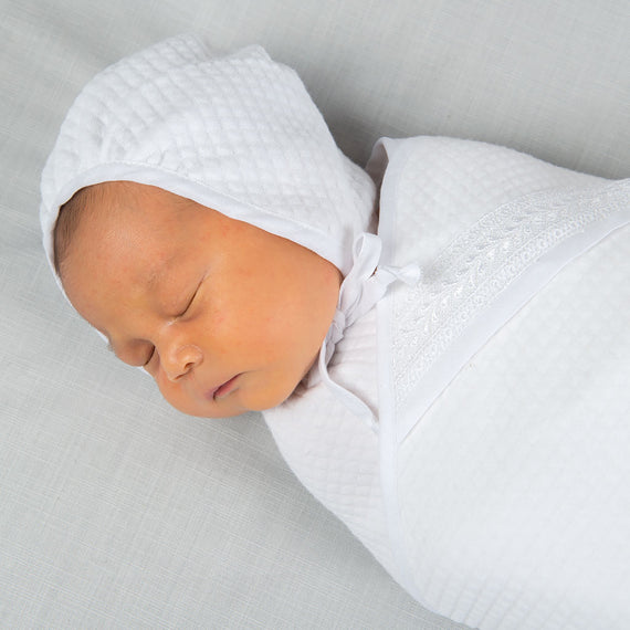 Newborn baby wearing an Elijah Newborn Quilted Bonnet made with soft quilted cotton in white and trimmed in white silk. White linen ties under the chin of the baby