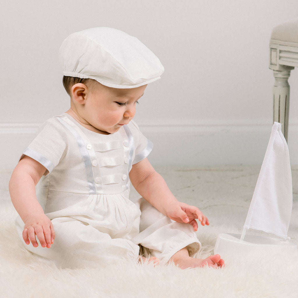 Baby boy sitting down and wearing the Owen Newsboy Cap and matching Linen Romper featuring silk ribbon in ivory and light blue across the front bodice and sleeves