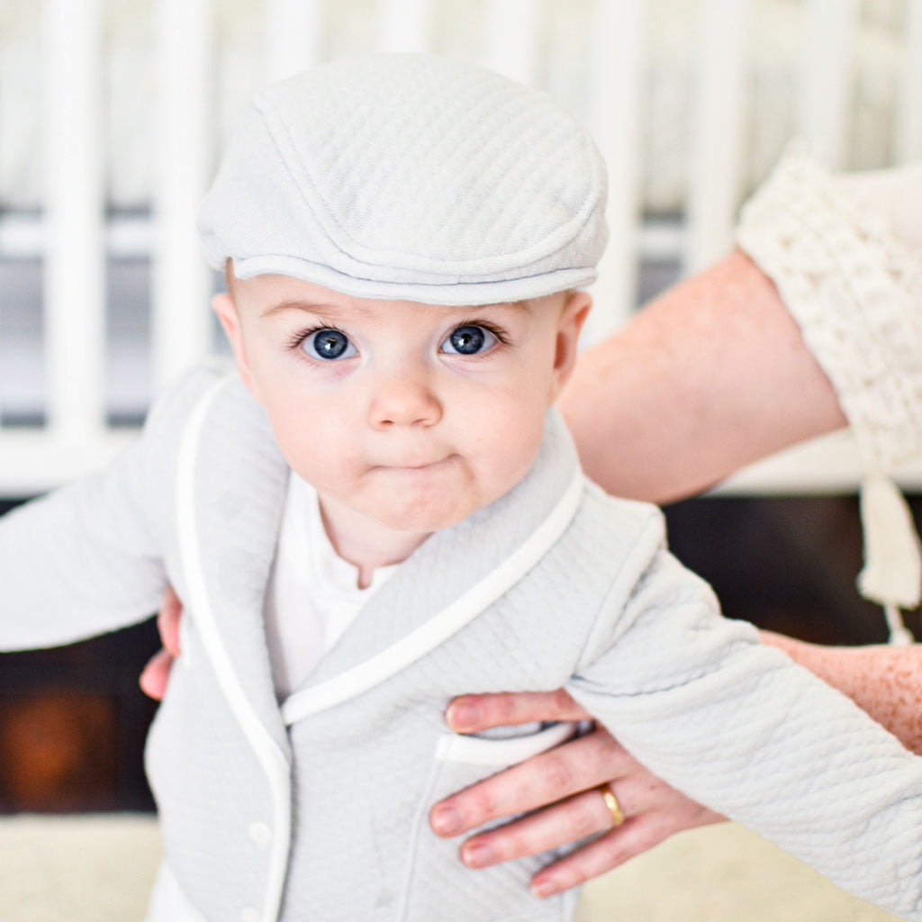 Baby boy smiling wearing gray newsboy cap. His mother is holding him by the arms.