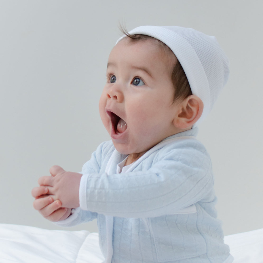 Baby boy sitting on the ground and wearing the Logan 3-Piece Suit (and White Ribbed Cotton Beanie), including the jacket, pants and onesie. Jacket made from blue textured cotton with a white French terry cotton trim.