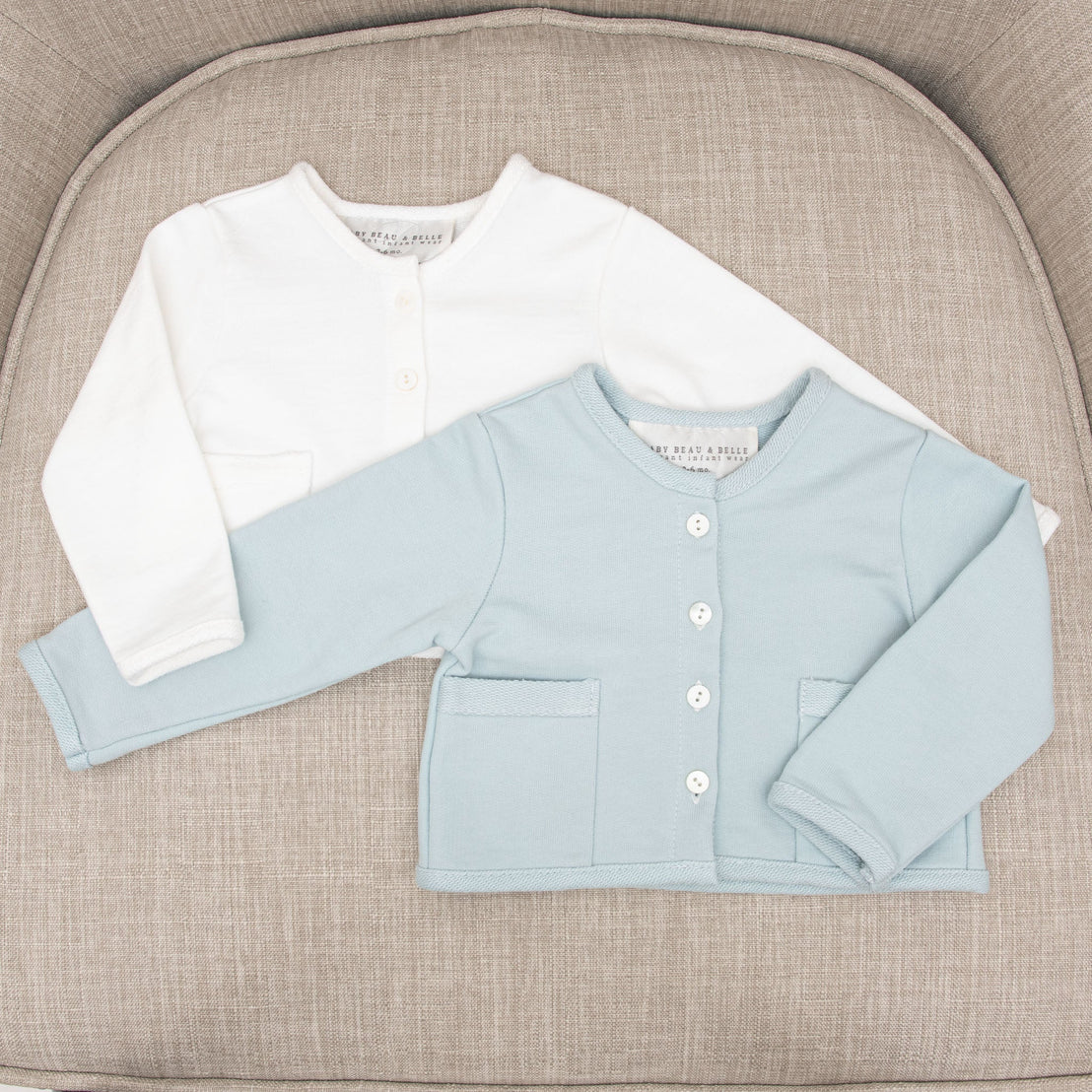 Two Isla French Terry Jackets, one white and one light blue are displayed on a beige textured cushion. Both jackets feature buttons and a pocket detail.