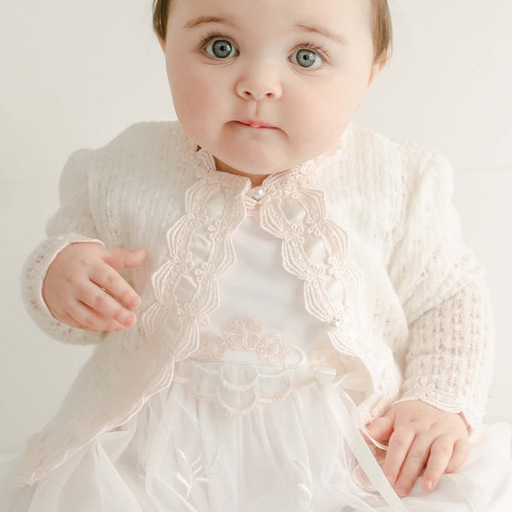 A baby with big blue eyes wearing a Joli Knit Sweater & Bonnet looks curiously at the camera, against a light background.