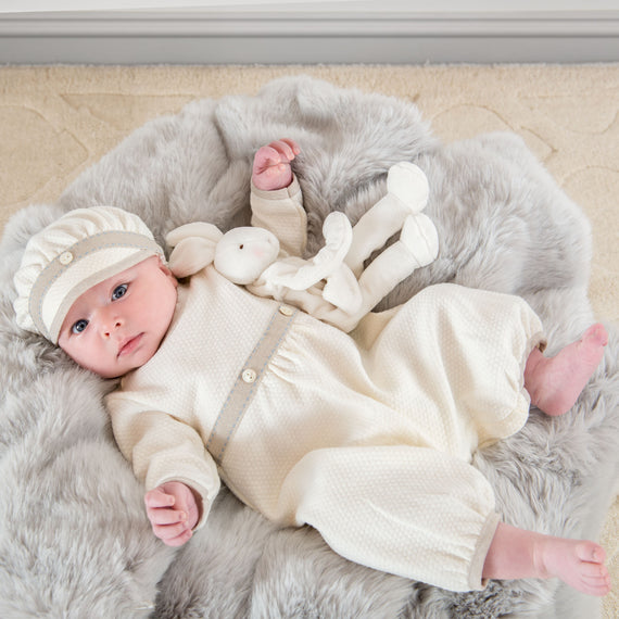 A baby dressed in an Austin Romper and striped hat lying on a gray furry blanket with a soft toy bunny, looking at the camera with a curious expression.