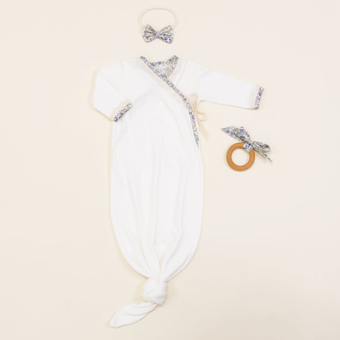 A vintage-inspired Petite Fleur Knot Gown with matching floral prints on the cuffs and neckline, paired with a headband and a boutique wooden teething ring, displayed on a soft beige background.