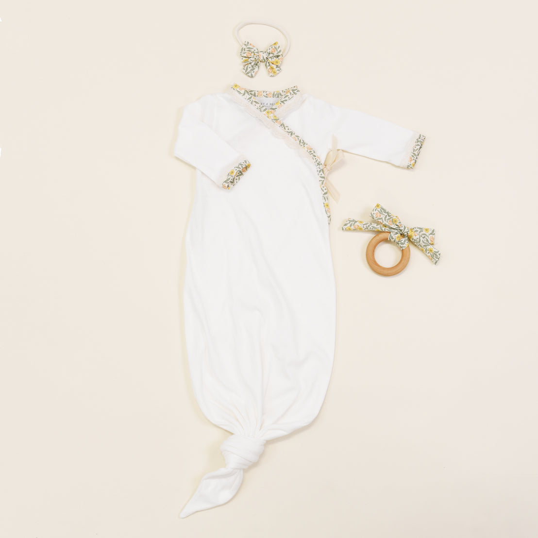 A Petite Fleur Knot Gown with decorative silver and gold sequined trim, accompanied by a matching bow headband and a wooden teething ring, displayed on a light beige background. This boutique set features vintage