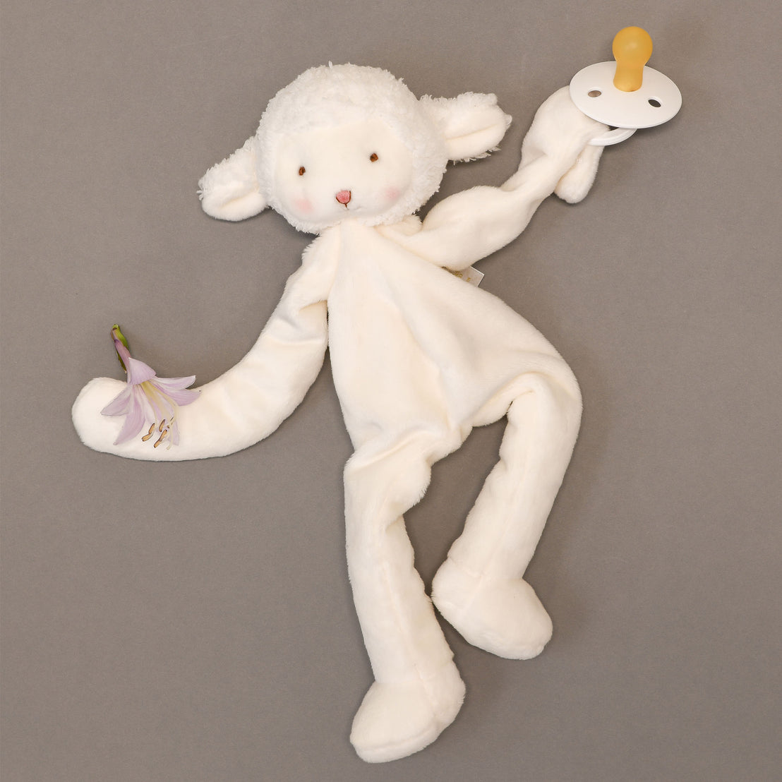 Introducing the Silly Lamb Buddy & Pacifier, a charming plush lamb toy in white with extended limbs clutching a purple flower and pacifier. Its soft, fuzzy texture and gentle expression make it the perfect christening gift, showcased against a simple gray background.