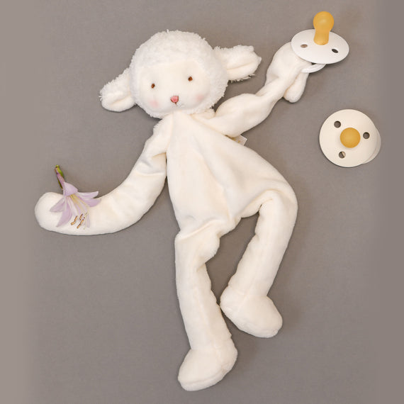 The Silly Lamb Buddy & Pacifier, with its long limbs, rests on a gray surface, cuddling a purple flower in one paw and holding a pacifier in the other. Ideal for a christening gift, it includes an attached pacifier clip for extra convenience.